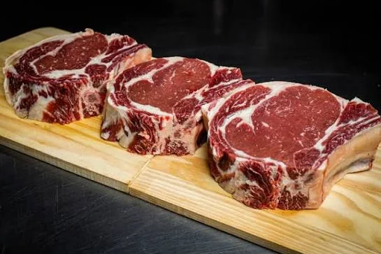 Três bifes ancho maturados sobre tábua de madeira, marmoreio evidente.